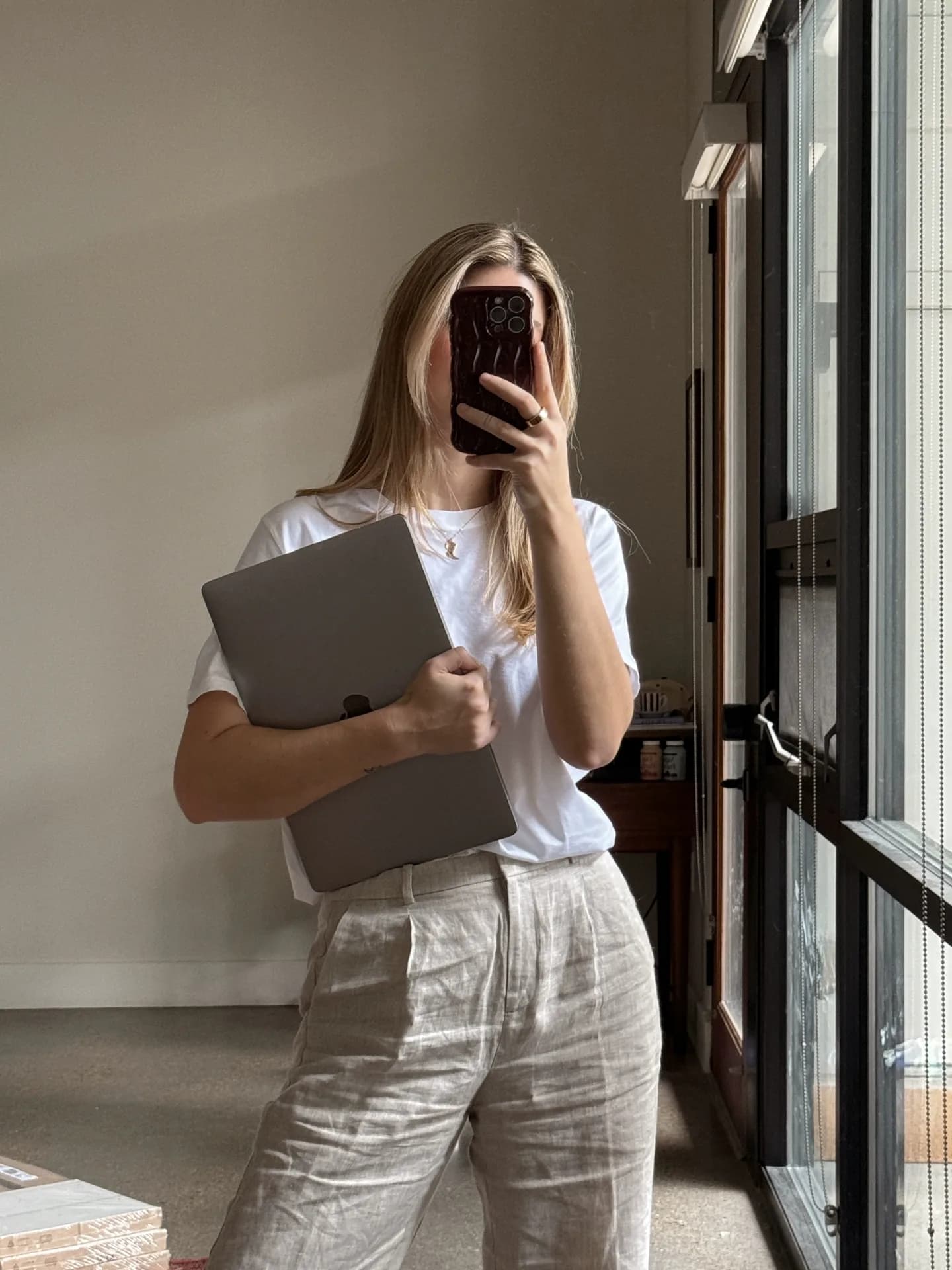 Žena u svetloj odeći fotografiše se telefonom u ogledalu dok u ruci drži laptop, pored velikog prozora kroz koji dopire dnevna svetlost.
